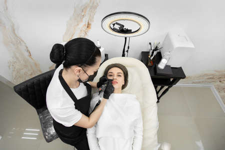 Cosmetologist wearing a mask and gloves applies permanent makeup to the lips of a young girlの写真素材