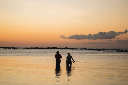 Two men fishing in the ocean from the beach at sunset.の写真素材