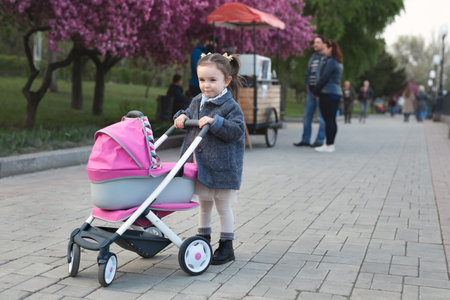 Little girl in a coat walks in the park and rolls a toy stroller with a doll