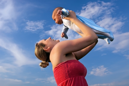Happy young mother lifting her baby boy high up against blue sky background の写真素材