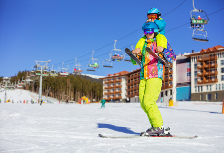 Father carrying tired son in skiing outfit on shoulders. Standing on ski slope with chairlifts.の写真素材