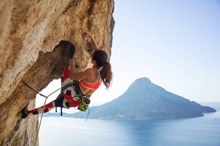 Young woman struggling to climb ledge on cliff, against view of sea and islandの写真素材