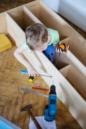 Cute young boy using screwdriver while sitting on floor at unfinished shelf unit or bookcase and holding tape measure in another handの写真素材
