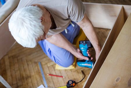 Mature man using electric screwdriver while making bookcase or shelf unit, view from aboveの写真素材