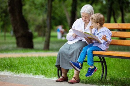 Young boy and great grandmother reading book in summer park, grandson eating bunの写真素材