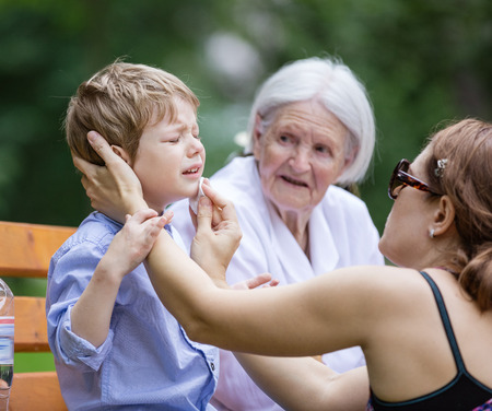Mother treating scrape on son's chin while sitting on bench in park. Senior grandmother sitting beside on bench.の写真素材