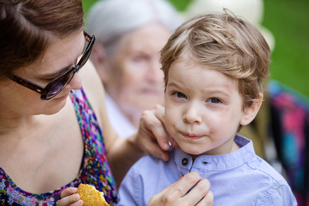 Mother comforting son after falling down and hurting face during walk in park. Boy eating cookie, senior grandmother sitting beside on bench.の写真素材