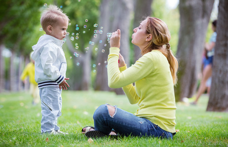 Mother blowing soap bubbles outdoors, little son watching her. Family having fun in park.