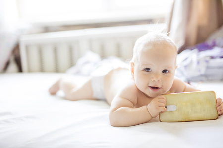 Cute baby girl with smartphone looking at camera while lying down on bedの写真素材