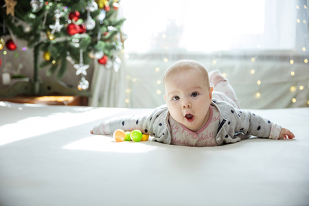 Cute baby girl lying down on bed at home. Christmas tree in background. Girl looks surprised and emotional.の写真素材
