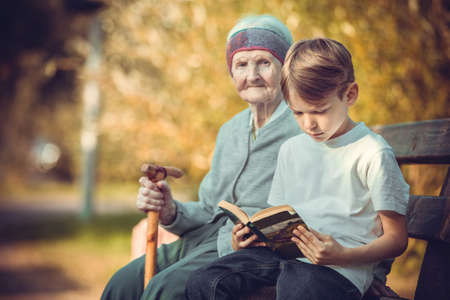 Young boy reading book on bench in park. His great grandmother sitting next to him.の写真素材