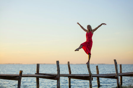 Beautiful young woman in a red dress posing on jetty at sunsetの写真素材