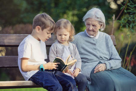 Young children and their great grandmother reading book on bench in parkの写真素材