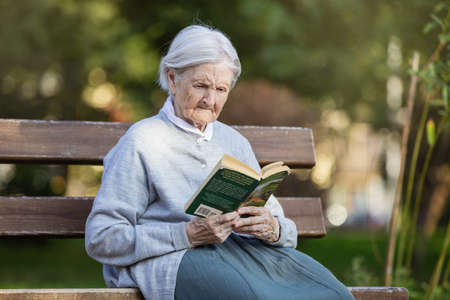 Portrait of senior woman on bench in park reading fiction bookの写真素材