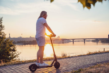 Caucasian man riding electric scooter in Kyiv at sunset. Evening view of Dnipro river from left bank.の写真素材