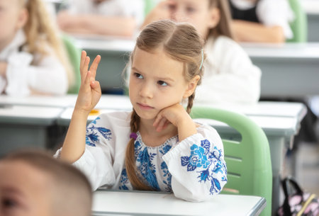 Cute young girl sitting at desk at school. The girl is raising her hand and showing four fingers as response to teacher's question. She is wearing traditional Ukrainian shirt.の写真素材
