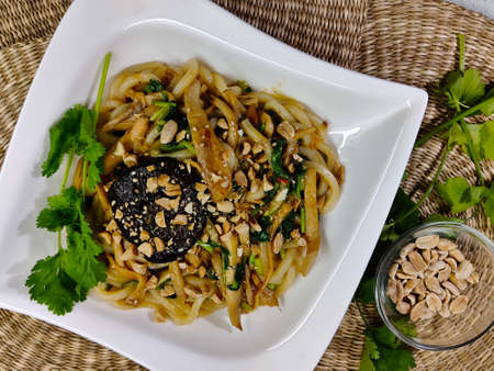 A dish made of potato stripes with parsley, peanuts, shiitake mushroom, and soy sauce served in white plate. There is parsley and bowl of peanuts on the left side of the image as well.の写真素材