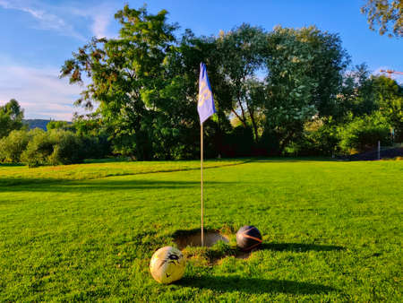 Marked hole with flag in the football golf field with two balls around it. It's played on nicely mown grass with trees in the background.の写真素材