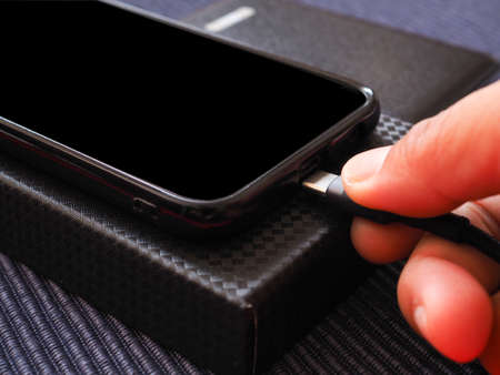 The hand is plugging the phone charger from the power bank on a black box over a blue background. Close up.の写真素材