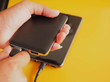 Close up woman's hand holding power bank is charging a smartphone on a yellow background. Copy space for text.の写真素材