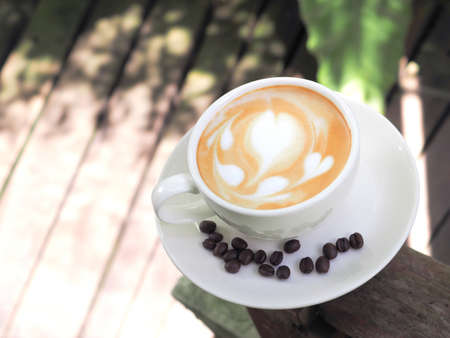 Hot coffee with heart shape, latte art in a white cup with coffee beans, on a wooden table in the garden, there is a comfortable atmosphere in the sunshine.の写真素材