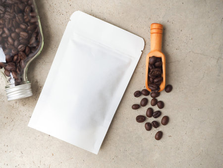 White paper bag with zipper and coffee beans in a bottle, wooden scoop on a grey background. Top view with copy space.の写真素材