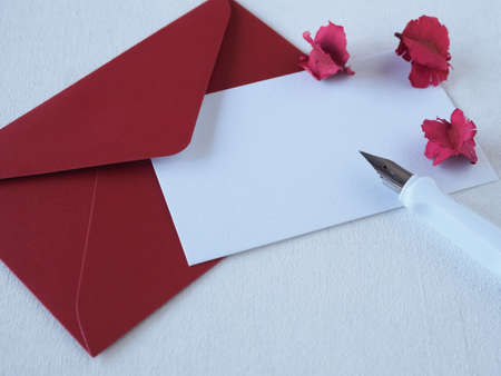 Pen on white paper with space for text and red envelope decorated with pink dried flowers over a white wooden background. Concept love, greeting card.の写真素材
