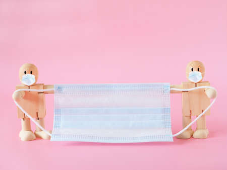 Two wooden dolls hold a medical face mask on a pink background. Concept of coronavirus outbreak and pollution. New normal.の写真素材