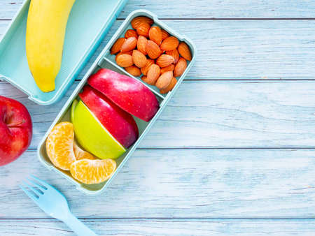 Fresh fruit in light green lunch box with almond, apple, orange, banana and fork on light blue wooden background. Take away food. Top view with copy space, healthy eating concepts.の写真素材