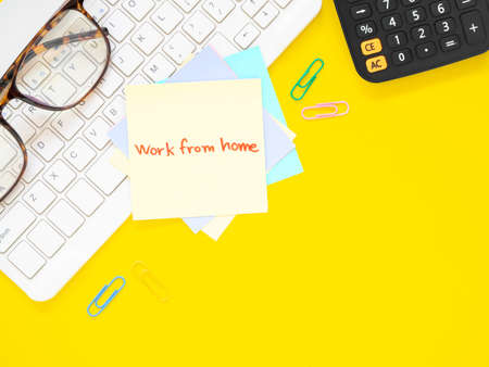 Yellow sticky paper Write a handwritten message "work from home" on keyboard and calculator, eyesglass, over yellow workplace desk table. Top view with space for text. Flat lay, Business and education concept.の写真素材