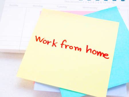 Yellow sticky paper Write a handwritten message "work from home" on monthly notebook over white wooden background.の写真素材