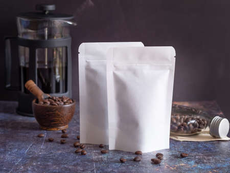 Roasted coffee beans in white paper bag with zipper and wooden bowl, glass bottle, coffee pot on grunge background. Space for your text.の写真素材