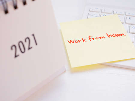 Yellow sticky paper Write a handwritten message "work from home" on the keyboard over white wooden background. The foreground of the calendar 2021 is blurred.の写真素材