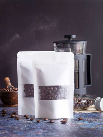 Roasted coffee beans in white transparent window paper bag with zipper and wooden bowl, glass bottle, coffee pot on grunge background. Space for your text.の写真素材