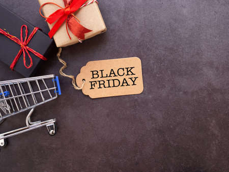 Top view, brown tag with text Black Friday and shopping cart, gift box on dark grey background. Flat...