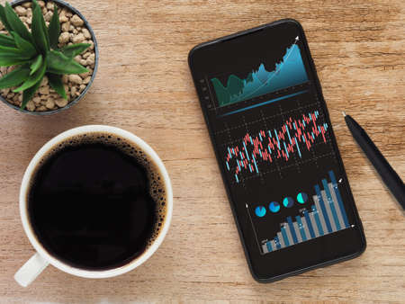 Top view, Smartphone show forex trading graph financial data on device screen with coffee cup and pen on wooden table. Online Investment and stock trading concept. Business finance background.の写真素材