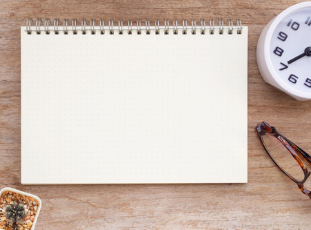 Top view, blank notebook and white clock show time 7 o' clock, eyeglasses and cactus on wooden table. Flat lay, space for your text.の写真素材