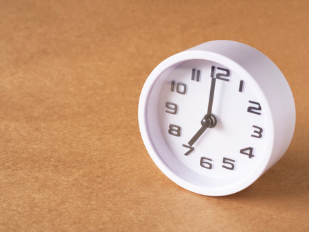 Close up, white clock shows time at 7 o'clock on the office brown workplace tabletop. Copy space for...