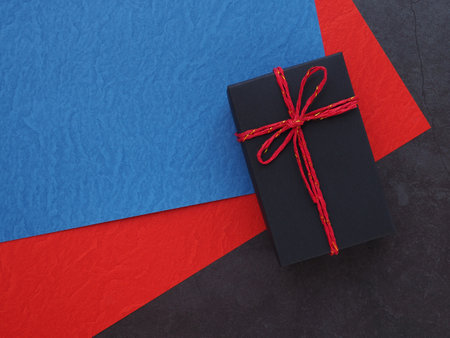 Black gift box tie red rope on blue, red paper over dark grey background. Holiday season concept. Top view, flat lay with copy space.の写真素材