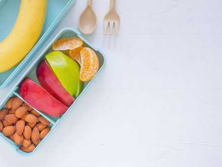 Top view, light green lunch box of fresh fruit with apple, banana, orange, almond and spoon fork on white wooden background. Take away food. Top view with copy space, healthy eating concepts.の写真素材