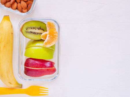 Top view, lunch box of fresh fruit with apple, kiwi, banana, orange and almond, fork on white wooden background. Take away food, with blank space for text, healthy eating concepts.の写真素材