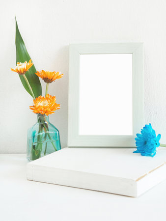 White wooden rectangle photo frame and orange flower, blue (chrysanthemum) with green leaves in vase...