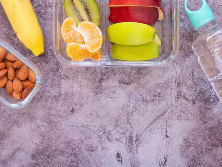 Glass container lunch box fresh fruit with apple, kiwi, banana, orange and almond, water bottle on grey background. Take away food. Top view with copy space, healthy eating concepts.の写真素材