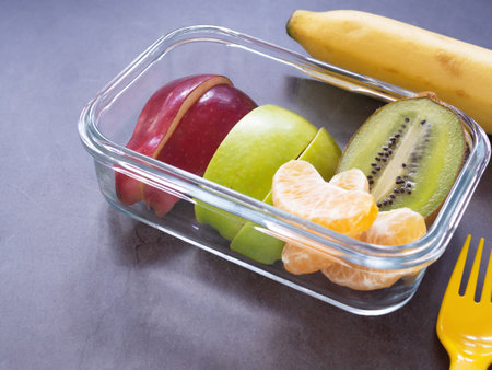 Fresh fruit in lunch box with apple, orange, kiwi and banana, fork on grey background. Take away food. Copy space with text, healthy eating concepts.の写真素材