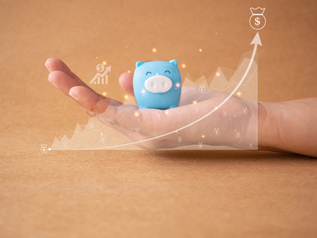 Savings concept. Hand holding blue piggy bank and graph growing with money bag dollar icon on brown background. Future financial stabilityの写真素材