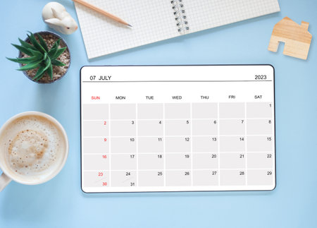 Top view, tablet computer showing July 2023 calendar app page and cactus, coffee cup, pencil, a bunny statue on blue background. Calendar application for schedule planning.の写真素材