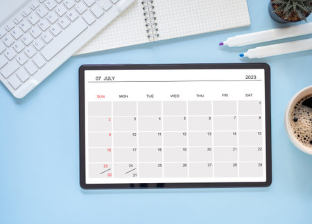 The online calendar app on the tablet computer showing screen July 2023, on blue background and keyboard, notebook, cactus. Reminder with the calendar application for schedule planning. Top view.の写真素材