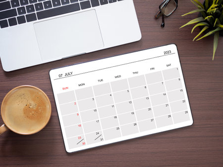 The online calendar app on tablet computer showing screen July 2023, on wooden workplace and coffee cup, laptop. Reminder with the calendar application for schedule planning. Top view, flat layの写真素材
