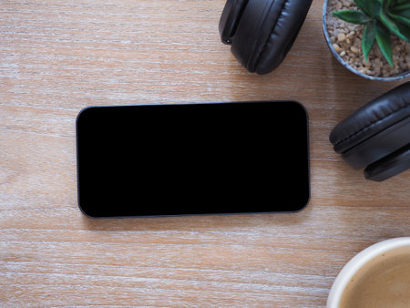 Top view, blank black screen of smartphone and cactus, headset, coffee cup on wooden background. Template, mock-up empty screen of mobile phone for text and logo of product.の写真素材