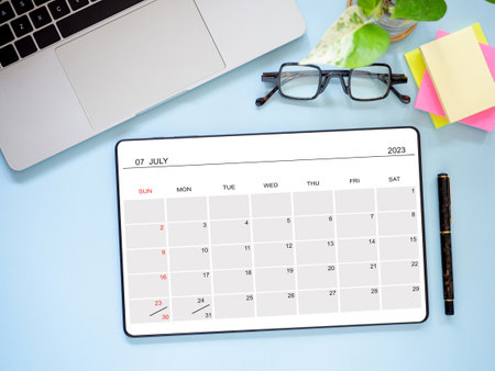 Top view, flat lay tablet computer showing July 2023 calendar app page and laptop, pen, and...